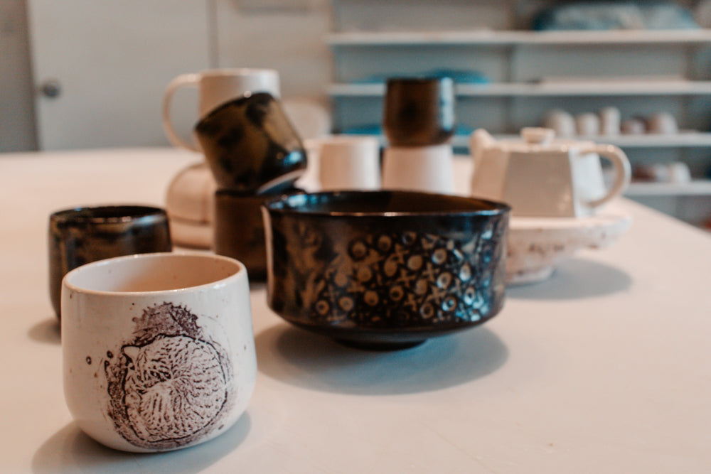 Une pièce de poterie avec un dessin de chat en avant plan se trouve sur une table. Dans le fond se trouvent d'autres pièces de poterie foncées qui sont empilées.