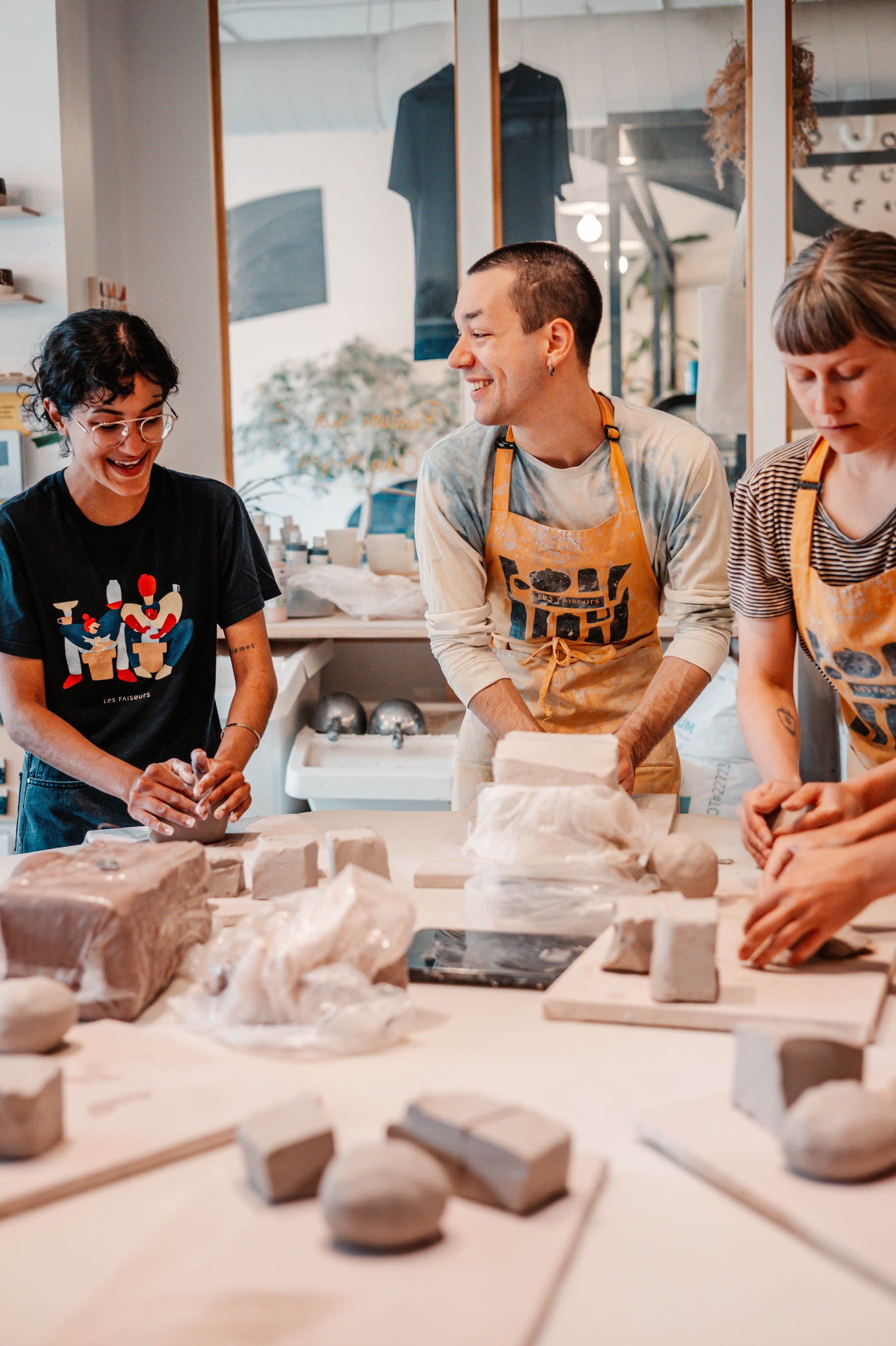 Trois personnes apprennent à faire de la poterie en s'amusant. Elles portent des t-shirts et tabliers Les Faiseurs